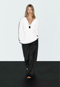 Hvid oversized bluse med en dyb V-hals, parret med løse sorte bukser. Enkel sort halskæde fremhæver outfittet. Glatte teksturer ses.