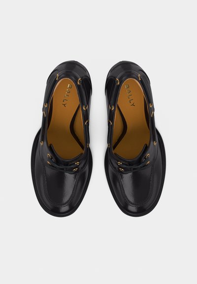 Ζευγάρι μαύρα γυαλιστερά δερμάτινα παπούτσια με κορδόνια, με χρυσές τρύπες και εσωτερικά ταμπά πέλματα με την επωνυμία "BALLY", φαίνονται από πάνω σε λευκό φόντο.