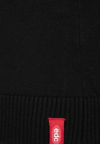 Schwarzer Strickpullover mit geripptem Saum und einem rechteckigen roten Etikett mit dem "edc"-Logo am unteren Rand. Weiche Textur mit glatter Oberfläche.