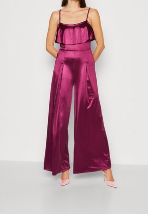 Kvinde iført en ærmeløs jumpsuit i magentarød satin med vide ben og flæsedetalje, parret med lyserøde højhælede sko.