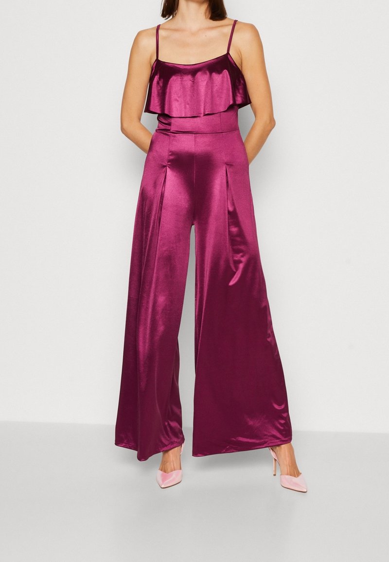 Femme portant une combinaison sans manches en satin magenta avec pantalon à jambes larges et détail volants, associée à des chaussures à talons hauts rose pâle.