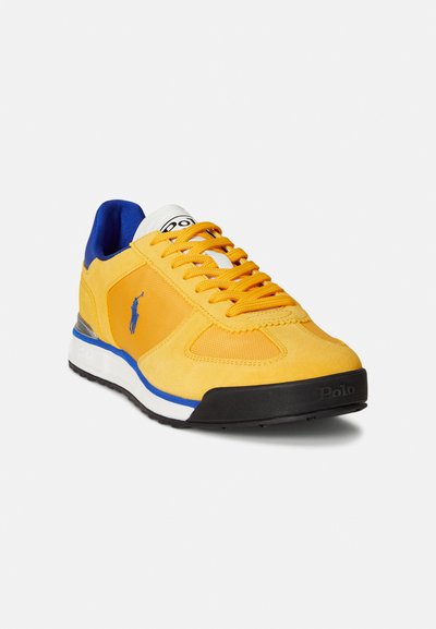 Żółty but sportowy wykonany z zamszu i siateczki, z niebieskimi akcentami, teksturowanymi sznurówkami i czarną podeszwą. Posiada logo na boku i języku.