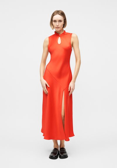 Γυναίκα που φοράει φωτεινό κόκκινο μεσομήκι αμάνικο φόρεμα με άνοιγμα στην περιοχή του λαιμού και σκίσιμο μπροστά, σε συνδυασμό με μαύρα παπούτσια χωρίς κορδόνια.