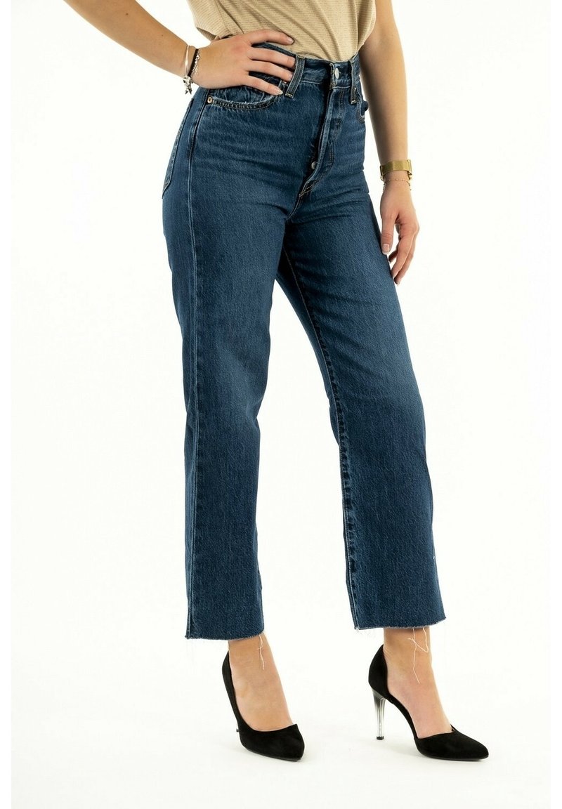 Jean en denim bleu taille haute avec une coupe droite, équipé d'une fermeture éclair, de cinq poches et d'ourlets effilochés, associés à des talons hauts noirs.