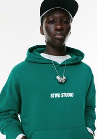 Ung person iført en mørkegrøn hættetrøje med teksten "STWD STUDIO", et sølvfarvet chunky halskæde, en hvid skjorte under og en sort baseballkasket.