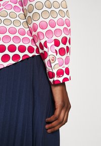 Bomullsskjorta har en vit bakgrund med stora rosa, röda och bruna prickar. Den har en knappad manschett och en lager på lager, marinblå, plisserad kjol.