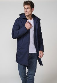 Temno modra parka z zadrgo in gumbom, ki ima kapuco in stranske žepe. Tkanina se zdi gladka in odporna proti vremenskim vplivom.