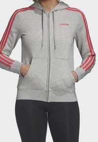 Felpa grigia Adidas con zip, featuring dettagli a tre strisce rosse lungo le maniche, tasche frontali e cappuccio con cordino. Tessuto morbido.