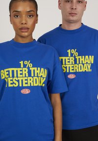 Tricourile din bumbac albastre au un text îndrăzneț de culoare galbenă care spune „1% MAI BUN DECÂT IERI.” Un logo este vizibil aproape de tivul fiecărui tricou.