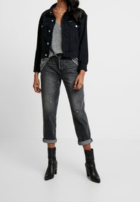 Schwarze Jeansjacke mit silbernen Knöpfen, graues V-Ausschnitt-Top, ausgefranste schwarze Jeans und schwarze Stiefeletten mit spitzem Zeh und Absatz.