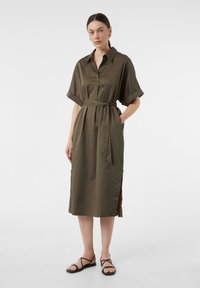 Olivgrünes Hemdkleid mit Kragen, kurzen umgeschlagenen Ärmeln, tailliert mit einem Gürtel und seitlichen Schlitzen. Das Modell kombiniert es mit schwarzen Riemchensandalen.