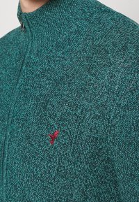 Grön stickad zip-sweater med hög krage, med ett texturerat mönster och en liten röd fågel-logotyp på bröstet.