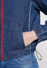 Denimjacka med dragkedja fram, röd dragkedja och vit piping. Har elastiska ärmmuddar och en ficka. Blå färg med en strukturerad finish.