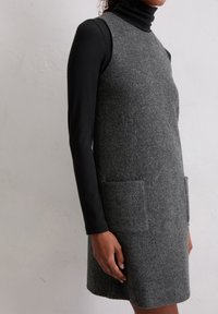 Grijs wollen jurk met hoge hals, mouwloos ontwerp, zijzakken en een structuurafwerking, gedragen over een zwarte lange mouwen shirt.