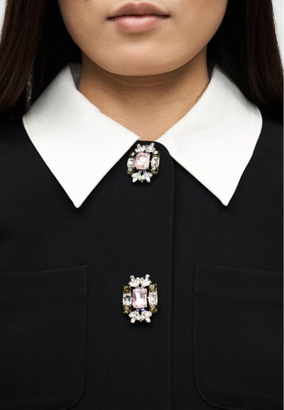 Γυναίκα που φοράει μαύρο ένδυμα με λευκό μυτερό γιακά και δύο διακοσμητικά κοσμημένα καρφίτσες, μία στον γιακά και μία στο στήθος.