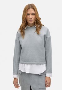 Sudadera gris claro con escote redondo, hombros caídos y dobladillo acanalado, superpuesta sobre una camisa blanca con cuello. Textura suave.