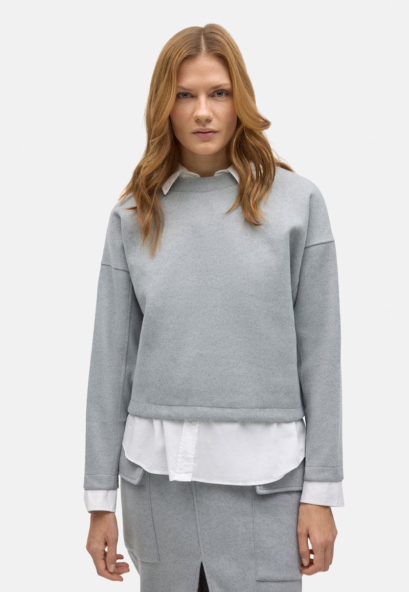 Sudadera gris claro con escote redondo, hombros caídos y dobladillo acanalado, superpuesta sobre una camisa blanca con cuello. Textura suave.