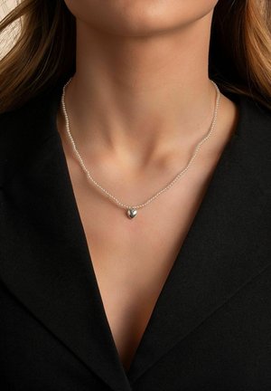 Pärlhalsband med dubbel rad, prytt med en liten hängande hjärtan i silver. Bärs tillsammans med en mörk blazer, vilket framhäver den delikata designen.