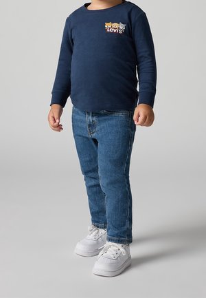 Småbarn iført en navyblå Levi's langærmet trøje med dyregrafik, blå jeans og hvide sneakers, der står på en neutral baggrund.