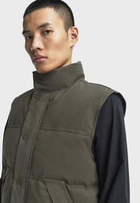 Olivgrüne gepolsterte Weste aus glattem Stoff, mit hohem Kragen, Druckknöpfen und Seitentaschen. Schwarzes Hemd darunter für den Lagenlook.