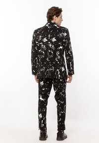 Mężczyzna stojący tyłem, ubrany w czarny garnitur z białym wzorem abstrakcyjnych plam farby i czarnymi butami na białym tle.