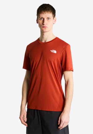 Jonge man draagt een kortärmlig shirt van The North Face in roestkleur en zwarte korte broek, staand tegen een effen lichte achtergrond.