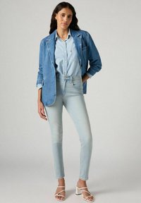 Blazer de mezclilla azul claro sobre una camisa de botones azul claro, combinado con jeans ajustados azul pálido y sandalias de tacón alto blancas con tiras.