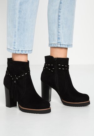 Bottines en daim noir à talons carrés, coutures blanches décoratives et pampilles, portées avec un jean court bleu clair sur un sol blanc.
