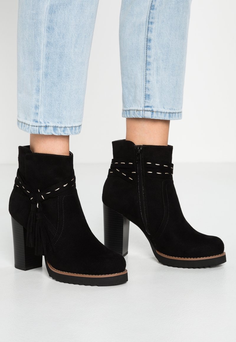 Bottines en daim noir à talons carrés, coutures blanches décoratives et pampilles, portées avec un jean court bleu clair sur un sol blanc.