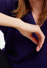 Femme à la peau claire et aux cheveux roux portant un pull violet, la main tendue avec les doigts légèrement courbés près de la poitrine.