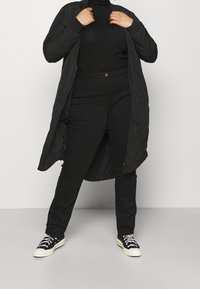 Person som bär en svart vadderad lång jacka, svart ribbad polotröja, svarta byxor och svarta sneakers med vita sulor och snörning.