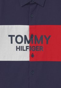 Granatowo-czerwona koszulka polo z białym poziomym paskiem. Na środku granatowy napis "TOMMY HILFIGER". Cztery czarne guziki na plisie.