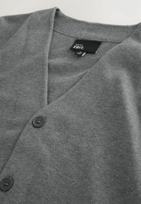 Cardigan gris doux boutonné avec col en V et étiquette noire indiquant "NEXT EDIT." Taille AA, fabriqué en Chine.