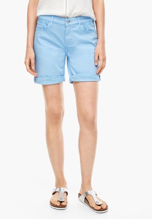 Person trägt hellblaue hochgekrempelte Shorts, weißes Top und Sandalen mit silberner Schnalle, steht auf weißem Hintergrund.