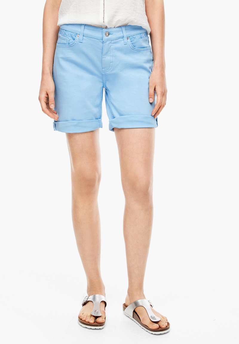 Personne portant un short bleu clair retroussé, un haut blanc et des sandales à boucle argentée, debout sur un fond blanc.