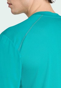 Turkos atletisk tröja med nätstruktur på baksidan, med en slät front och kontrasterande sömmar längs kanterna.