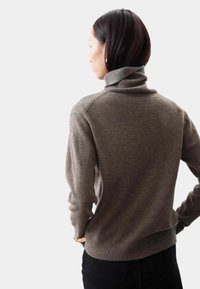 Brauner Rollkragenpullover mit geripptem Kragen und Bündchen, aus weichem Material. Getragen mit dunklen Hosen, die ein schlichtes, tailliertes Design zeigen.