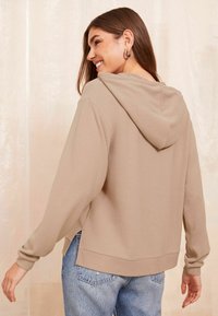 Cappuccio corto color khaki con cappuccio regolabile, polsini e orlo a costine, abbinato a jeans in denim chiaro. Tessuto morbido e vestibilità rilassata.