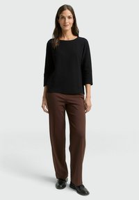 Schwarzes, dreiviertellanges Oberteil mit Rundhalsausschnitt, kombiniert mit braunen, weit geschnittenen Hosen. Beide Teile haben eine glatte Textur. Schwarze Schuhe.