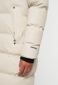 Lys beige pufferjakke med en glat tekstur, der har en lynlåslomme og et sort logo på ærmet. Manchetterne er sorte.