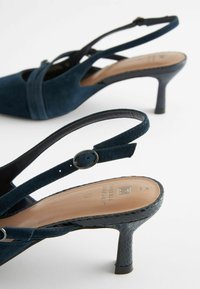 Navy blue suede slingback heels with a pointed toe, slender heel, and crisscross strap design. Textured heel accent adds detail.