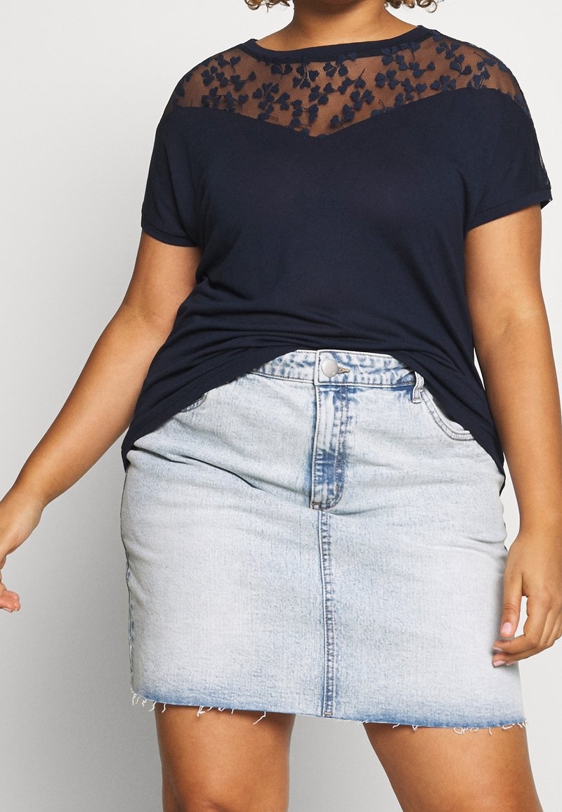 Marinblå t-shirt med blommönstrad meshnätöverdrag vid bröstet; kombinerad med en ljusblå denim mini kjol med fransad fåll och knappstängning fram.