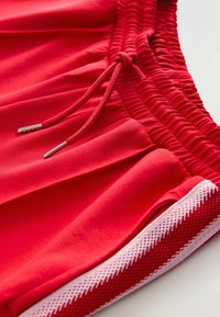 Shorts de sport rouges en tissu lisse, dotés d'une taille élastique avec cordon de serrage ajustable et d'accents en maille sur les côtés.