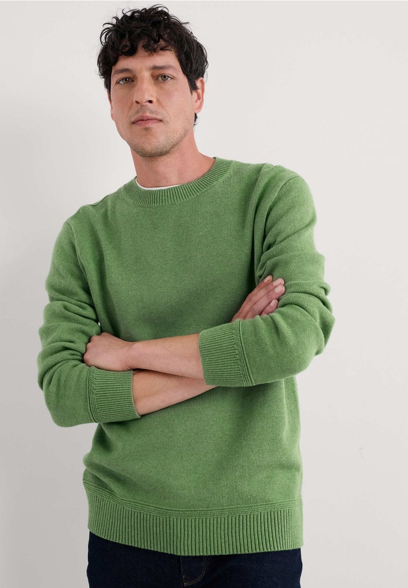 Suéter verde de punto con un escote redondo, puños y dobladillo acanalados, con un acabado texturizado. Llevado con jeans oscuros.