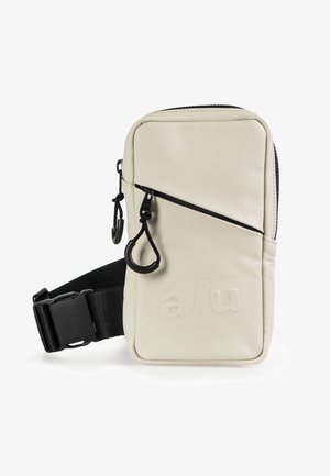 Kompakt beige crossbody taske med sort justerbar strop, diagonalt lynlås på frontlommen og prægede "a/u" logo på frontpanelet.
