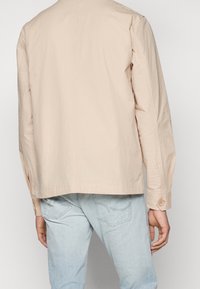 Chaqueta beige clara con un acabado texturizado, con un corte recto y detalles de puños abotonados, llevada sobre unos vaqueros azul claros.