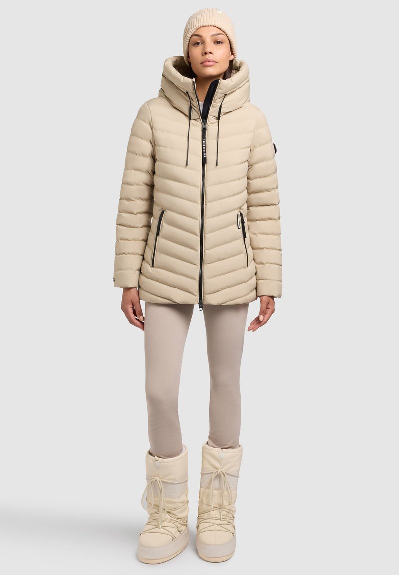 Giacca puffer beige con cappuccio, chiusura con zip e trapuntatura orizzontale. Abbinata a stivali da neve color crema e leggings beige.