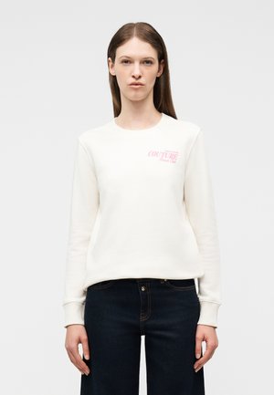 Ženska, oblečena v belo majico z dolgimi rokavi z roza napisom "Couture Beach Club" na prsih in temno modrimi kavbojkami, stoji ob belem enobarvnem ozadju.