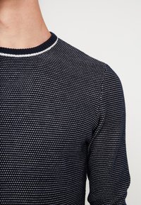 Maglione lavorato a maglia di colore blu navy con un motivo strutturato e un accento bianco sul collo. Presenta un colletto rotondo e maniche aderenti.