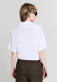 Camicetta bianca a maniche corte con colletto, texture liscia. Abbinata a pantaloni elasticizzati marroni scuri. Vista dal retro.